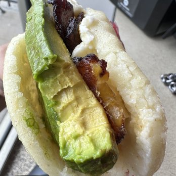 Arepa Queen - Arepa tropical - A detailed photo of a Venezuelan arepa with avocado slices, crispy bacon, and melted cheese, held outdoors near a building. - Venezuelan Near Me - Frederick, Maryland