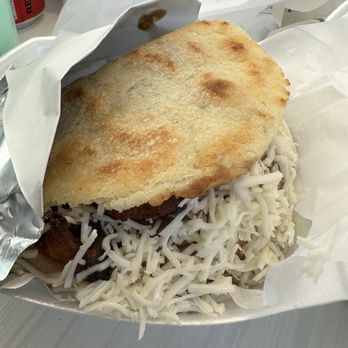 Arepasmanía - a close up of a plate of food - A food dish in a paper container includes shredded cheese, a fried plantain, and a piece of meat underneath. The fried arepa is placed on top, partially covering the other ingredients. The container is lined with white paper, and the background shows a table with some cups and wrappers. The dish appears to be a typical Latin American or Venezuelan meal. - Venezuelan Near Me - Miami, Florida
