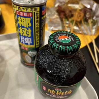 Mala Siblings - A can of coconut juice with a pull-tab opening is positioned behind a black bottle of Chinese herbal medicine on a tray. The can features bright yellow and blue Chinese characters, and the bottle has a black cap with green lettering. In the background, there are blurred images of skewered food and people at a dining setting. - Specialty Food Near Me - Flushing, New York