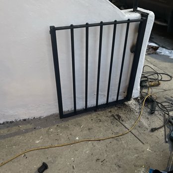 JK Welding - A black metal gate with vertical bars is propped against a white exterior wall, with tools and extension cords scattered on the concrete ground. - Metal Fabricators Near Me - Brooklyn, New York