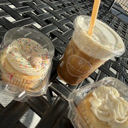 Cloud 9 ice coffee, vanilla cupcake and buttercream vanilla cupcake. - Two cupcakes with frosting and sprinkles, and an iced coffee in a clear cup with a straw, are placed on a black outdoor table.