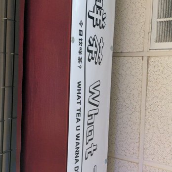 What Tea - Store sign - The image shows a rectangular sign mounted on a wall, featuring Chinese characters and the English phrase "What tea wanna drink?". The sign includes a cartoon cloud illustration at the top and is attached with visible screws. The background wall has a textured surface, and part of a vent or grille is visible on the right side. - Bubble Tea Near Me - Boston, Massachusetts