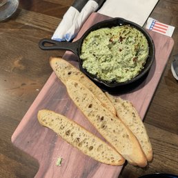 Ricotta dip..... - A serving of green dip in a cast iron skillet accompanied by three slices of toasted bread on a wooden board, set on a restaurant table.