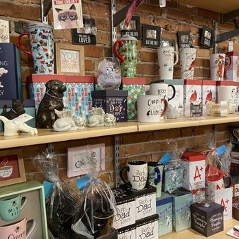 Dimple's - Arrangement of gift items including ceramic mugs with humorous and sentimental messages, small animal figurines, framed artwork, and decorative boxes on wooden shelves. The background is a brick wall, and the items are organized in a retail setting, likely a gift shop specializing in home decor and personalized gifts. - Women's Clothing Near Me - Racine, Wisconsin