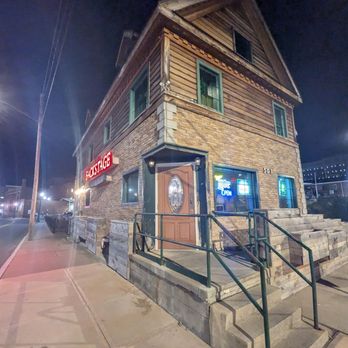 Backstage Pub - A two-story building with brick and wood siding, illuminated by neon signs, located on a sidewalk at night. The building has a staircase and a ramp at the entrance. - Burgers Near Me - Schenectady, New York