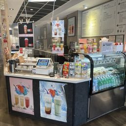 A tea shop counter with menu boards, promotional posters for drinks, and a refrigerated display case with bottled beverages, set in a cozy interior.