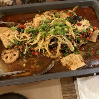 Too Hot - Grilled Whole Sea Bass with Spicy Ginger and Tofu - A large tray contains a spicy Asian-style fish dish topped with chopped green onions, shredded vegetables, and chili peppers. The dish includes tofu, lotus root slices, and other vegetables in a reddish-brown broth. In the background, there are small bowls of rice and empty dishes on a wooden table, indicating a shared meal setting. - Asian Fusion Near Me - Cambridge, Massachusetts