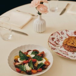 A close-up of a vibrant Caprese salad served in a white textured bowl next to a gourmet tartare dish on a cream-colored tablecloth.