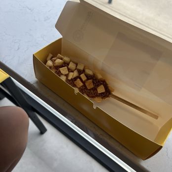 Mochinut - A yellow rectangular box with a clear plastic lid is open, revealing a dessert topped with small square pieces, possibly fruit or candy. The box is on a white marble-like table near a window, with a chair leg visible in the background. - Hot Dogs Near Me - Columbus, Ohio
