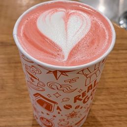 Cherry Blossom - A paper cup containing a latte with pinkish foam and white heart-shaped latte art on top is placed on a wooden table. The cup features decorative graphics and text related to coffee. The background shows part of a metal sink or counter area, indicating a café setting.