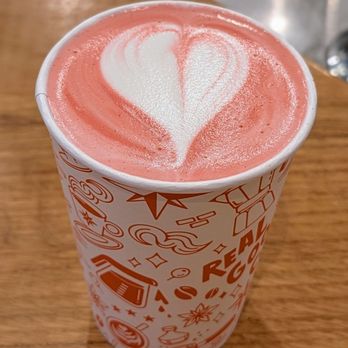 Compass Coffee - Cherry Blossom Latte - A paper cup containing a latte with pinkish foam and white heart-shaped latte art on top is placed on a wooden table. The cup features decorative graphics and text related to coffee. The background shows part of a metal sink or counter area, indicating a café setting. - Coffee Roasteries Near Me - Washington, DC, District of Columbia