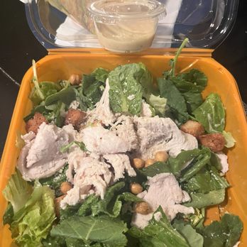 Wildflower - So underwhelming - A salad featuring leafy greens, sliced cooked chicken, chickpeas, and croutons is presented in an orange plastic container. A small cup of dressing is placed behind the salad, with a clear lid partially open. The salad appears fresh, with various types of lettuce and toppings, suitable for a casual dining or takeout setting. - Sandwiches Near Me - Tempe, Arizona