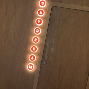 Daydream - A neon sign with the word "DAYDREAM" in red letters is mounted vertically on a wooden wall. The sign is illuminated and positioned next to a door with a metallic handle and a lock. The background consists of wood paneling, and the lighting emphasizes the glow of the neon sign. - Cocktail Bars Near Me - Albuquerque, New Mexico