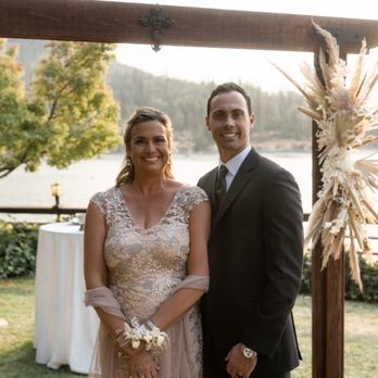 Light and Well - A woman in a lace dress and a man in a dark suit stand together under a wooden wedding arch decorated with dried floral arrangements. The background features a lake, trees, and a railing, with soft natural lighting indicating late afternoon or early evening. The scene is set outdoors with greenery and water visible behind them. - Floral Designers Near Me