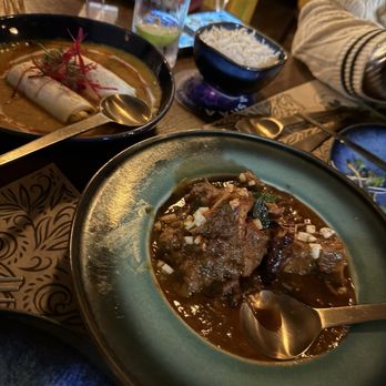 Tamashaa - GOAT VINDALOO and paneer pinwheel - Two Indian dishes in black bowls on a restaurant table, with glasses of water and a person in a striped sweater nearby. - Indian Near Me - Washington, DC, District of Columbia