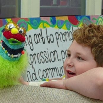 Create Community Studios - Puppet performance - A young boy with curly red hair rests his chin on his arm, looking at a puppet made of fuzzy, colorful materials with googly eyes and a yellow nose. The puppet is held up in front of a sign that reads '#g art to prom' and mentions community art activities. The background includes a window and a painted, colorful border, indicating an indoor community art studio setting. - Community Service/Non-Profit Near Me - Schenectady, New York