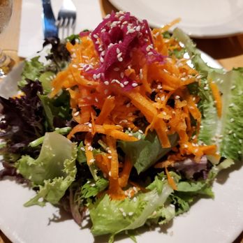 American Flatbread - A colorful mixed green salad features different types of lettuce, shredded carrots, and pickled vegetables, topped with sesame seeds. The salad is served on a white plate, with a fork and a glass of water nearby, on a wooden table. The presentation emphasizes fresh ingredients and vibrant colors. - Pizza Near Me - Rockport, Maine
