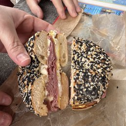 Ham egg cheese on the ET - A bagel sandwich with ham, egg, and cheese, cut in half, topped with sesame and black seeds, placed on parchment paper on a table.
