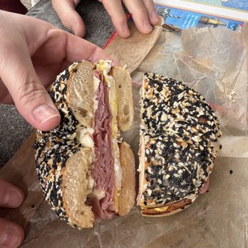 Schmagel's Bagels - Ham egg cheese on the ET - A bagel sandwich with ham, egg, and cheese, cut in half, topped with sesame and black seeds, placed on parchment paper on a table. - Bagels Near Me - Saint Augustine, Florida