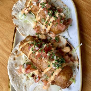 Casa del Cabo - Fish tacos - Two fish tacos are presented on a white oval plate, each with a fried fish fillet inside a soft tortilla, topped with shredded lettuce, diced tomatoes, chopped chives, and a drizzle of sauce. The plate is placed on a wooden surface, and the tacos are garnished with additional chopped chives and sauce. - Seafood Near Me - Eastham, Massachusetts