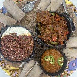 A plate of Ethiopian food features injera bread arranged around three small bowls containing different dishes: a minced meat stew, a vegetable stew with peppers, and a lentil-based dish garnished with green chili slices. The food is presented on a colorful, patterned tray with several rolled injera pieces and metal utensils. The setting suggests a traditional Ethiopian meal presentation.