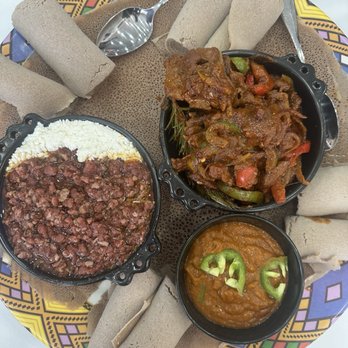 Habesha Cafe - A plate of Ethiopian food features injera bread arranged around three small bowls containing different dishes: a minced meat stew, a vegetable stew with peppers, and a lentil-based dish garnished with green chili slices. The food is presented on a colorful, patterned tray with several rolled injera pieces and metal utensils. The setting suggests a traditional Ethiopian meal presentation. - Desserts Near Me - Euless, Texas