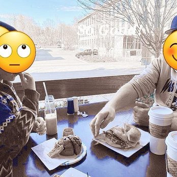 Boston Bean House - Two individuals with emoji faces sit at a cafe table near a large window, eating sandwiches and drinking coffee or milkshakes. - Coffee & Tea Near Me - Maynard, Massachusetts