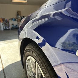 This is after the dent was fixed. - A blue vehicle's front fender and tire are visible, with a garage interior in the background, showing shelves and storage items.