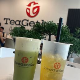 Thai Green Tea - Two tall glasses of iced drinks, one green and one yellow, are on a white table at TeaGather in Grand Prairie, TX. Both drinks have black straws and are labeled with the TeaGather logo. The background features the TeaGather logo on the wall and some indoor plants, with a few customers visible in the seating area.