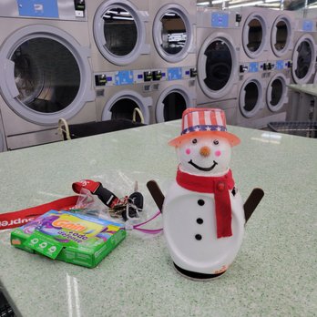 Laundry Palace - Folding tables - A snowman-shaped figurine with a red scarf and striped hat is placed on a laundry room table, with laundry machines in the background. - Laundry Services Near Me - Riverhead, New York