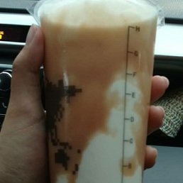 A person holds a large, clear plastic cup with a layered iced coffee beverage, showing distinct milk and coffee layers, inside a vehicle.