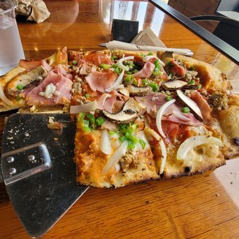 The Foundry Kitchen - Flatbread supremo/meat!! - A rectangular flatbread pizza topped with various ingredients such as ham, sausage, sliced mushrooms, onions, and chopped green onions. The pizza is placed on a wooden serving board, with a pizza cutter partially inserted into one corner. A glass of water is positioned to the upper left of the pizza, and the setting appears to be in a casual dining restaurant with a wooden table and natural lighting. - Salad Near Me - Springfield, Illinois