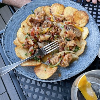 Quartiere - Veal - A round blue ceramic plate contains stir-fried vegetables and meat, with visible ingredients like onions, peppers, and minced meat, garnished with potato chips arranged around the perimeter. A fork rests on the plate, and a glass of water with lemon slices is partially visible in the foreground. The setting appears to be outdoors on a black metal table, with a person wearing a checkered shirt visible in the background. - Pizza Near Me - Stamford, Connecticut