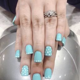 A close-up image of a person's hand displaying manicured nails painted in light blue with white polka dots. The hand is adorned with a diamond ring featuring multiple bands and a central diamond. The hand rests on a metallic surface, possibly a tray or table, with a blurred background. The nails are well-groomed and the ring is prominently visible.