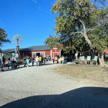 KC Pumpkin Patch - Outside - People are standing in a queue outside a red barn at a farm or pumpkin patch, with a large tree and a windmill nearby on a sunny day. - Pumpkin Patches Near Me - Olathe, Kansas