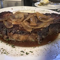 Prime Rib - A sizable prime rib steak is presented on a white plate, topped with caramelized onions. The steak appears cooked with a dark crust, and there is a sauce or juices around it. Some herbs are sprinkled on the plate for garnish. The background shows a restaurant setting with other plates and utensils.