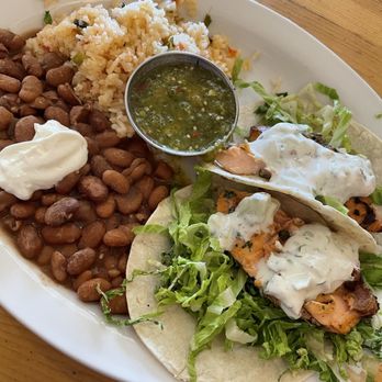 Tune-Up Cafe - Salmon Tacos - A white oval plate contains two salmon tacos with shredded lettuce and a white sauce. Accompanying the tacos are a portion of pinto beans with a dollop of sour cream, a serving of rice with vegetables, and a small cup of green salsa. The plate is placed on a wooden table, with a glass of dark beverage partially visible in the background. - New Mexican Cuisine Near Me - Santa Fe, New Mexico