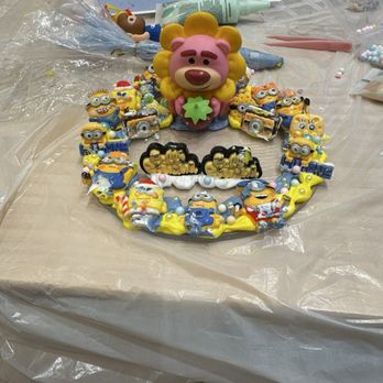 Hello Pottery - We had a good time spending at Hello Pottery. Our items turned out to look nice! We will be back again. - The image shows a Minions-themed cake topper with a pink lion figure holding a green star at the center. The topper is decorated with multiple Minions characters in various poses and expressions, arranged in a circular pattern around the lion. The table surface has baking tools, including piping tips, a tube of icing, and other decorating supplies, indicating a cake decorating activity. The scene appears to be set in a kitchen or bakery workspace. - Paint-Your-Own Pottery Near Me - Arcadia, California
