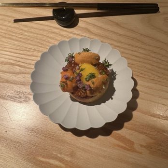 Ikigai - A food presentation featuring a small, round dish topped with garnishes, including herbs and edible flowers, on a white scalloped plate. Above the plate, a pair of chopsticks rests on a black chopstick holder on a wooden table surface. The setting suggests a fine dining experience, possibly at a Japanese or Kaiseki-style restaurant. - Japanese Near Me - Brooklyn, New York