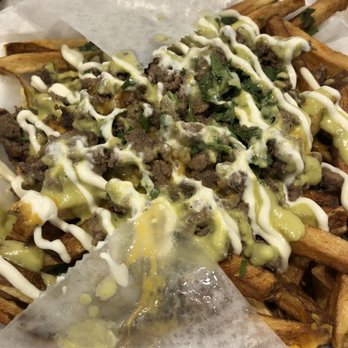 Dogos VIP - Carne asada fries - A takeout container holds carne asada fries topped with chopped herbs, drizzled with white and green sauces. The fries are visible around the edges, with some sauce spilling onto the container. The image shows a close-up of the dish, highlighting the toppings and sauces used. - Hot Dogs Near Me - Albuquerque, New Mexico