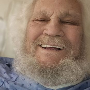 Blake's Lotaburger - A close-up image of an elderly man with white hair and a full white beard, smiling with eyes partially closed. He is wearing a blue hospital gown with a snowflake pattern, and the background suggests a hospital or medical environment. The man appears to be in good spirits, possibly resting or recovering. - Burgers Near Me - Moriarty, New Mexico