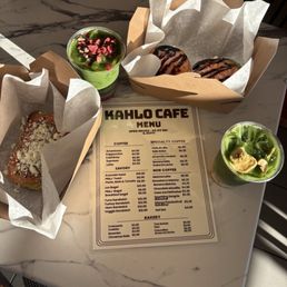 Chocolate Chip cookie and Brownie - A marble table holds two green drinks with toppings, two boxes of donuts, and a sandwich, with a Kahlo Cafe menu in the center.