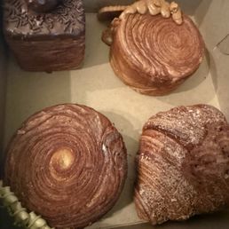 Rocher Croissant - The image shows four baked pastries inside a box. The top left pastry is a chocolate cake with chocolate sprinkles, a cherry, and gold leaf decoration. The top right and bottom left pastries are cinnamon rolls with a spiral shape and a glossy finish. The bottom right pastry is a croissant with a dusting of flour and a flaky, layered texture. The box appears to be a bakery box, and the pastries are arranged neatly.