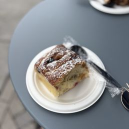Blueberry Babka - A slice of blueberry babka with a golden-brown crust and a soft, airy interior containing blueberry filling is served on a white disposable plate. The babka is dusted with powdered sugar and is accompanied by a black plastic knife wrapped in plastic. The plate is on a gray table, with another plate of food visible in the background, suggesting an outdoor dining setting.