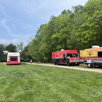 Civic Park - Food trucks - The image shows two food trucks parked side by side along a paved pathway in a park setting, with a large inflatable bounce house shaped like a dog on the grass nearby. The park has green grass, trees, and a few benches, with a person walking in the background. The sky is partly cloudy, and the scene appears to be during daytime. - Parks Near Me - O' Fallon, Missouri