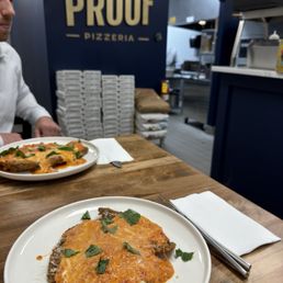 Lemon Zest Chicken Cutlet - Inside Third Proof Pizzeria in Pine Brook, NJ, two plates of chicken parmesan with melted cheese and herbs are placed on a wooden table. The restaurant's logo is visible on the wall in the background, along with stacked pizza boxes and kitchen equipment. A person in a white shirt is partially visible on the left side of the image.