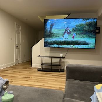 TV Geeks - Tricky install - mounted an 86 inch TV on a stairwell - The image depicts a living room with a large flat-screen TV mounted on the wall, displaying an animated scene from a cartoon. The room has light-colored walls, wooden flooring, and a gray sofa in the foreground. A small black table with a white device or speaker is positioned below the TV. The ceiling has recessed lighting, and a staircase with a black and white railing is visible in the background. The scene on the TV shows animated characters in a natural outdoor setting. - TV Mounting Near Me
