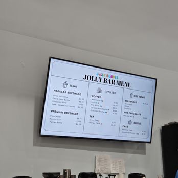 Jolly Bubble - Digital menu board at Jolly Bubble in Markham, ON, listing various drink options such as coffee, tea, milkshakes, and desserts. The menu includes categories like regular and premium beverages, with prices for items like Oasis Juice, caramel macchiato, rainbow cake, and hot chocolate. The display is mounted on a white wall, with some cups and utensils visible below the screen. - Kids Activities Near Me - Markham, Ontario