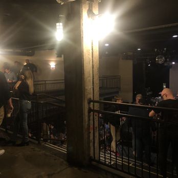 The Sinclair - View from the mezzanine after the lights came on - The image shows an indoor music venue with a stage on the right side, equipped with lighting and sound equipment. The venue has a mezzanine level with a railing, where some people are standing and observing. The lighting is dim, with bright stage lights and some wall-mounted fixtures. Several attendees are visible, some standing near the railing and others seated or standing further back. The space has an industrial style with exposed concrete columns and a dark ceiling. - Music Venues Near Me - Cambridge, Massachusetts