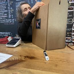 Sue packing up the half case of reds - A woman is handling a large cardboard box on a wooden table, with a chalkboard menu and photos in the background, in a cafe or shop setting.